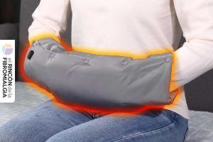 Dispositivo de soporte lumbar térmico para alivio del dolor en pacientes con fibromialgia, mostrando emisión de calor en tonos naranja y rojo, con una persona vestida con sudadera blanca y pantalones vaqueros sentada en un sofá gris, ilustrando el uso del producto. Aparatos para aliviar dolor de la fibromialgia y mejorar el día a día